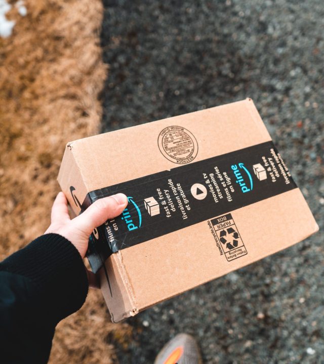 A person holding a cardboard Amazon Prime package on a snowy urban sidewalk.